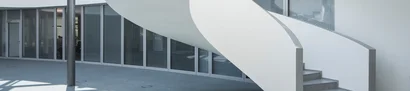 Innenansicht unseres GOB-Gebäudes: Das Atrium. Atrium mit geschwungener weißer Treppe, Glasfronten und grauem Boden im GOB-Gebäude.