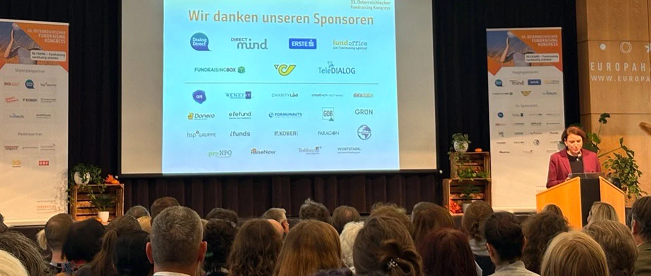 Begrüßung des Publikums beim ÖFK 2026 mit einer Rednerin am Pult und Sponsorenlogos auf der Leinwand.