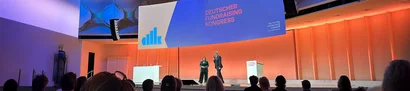 Deutscher Fundraising Kongress 2025: Eröffnungsbühne | 1905 x 425 Pixel Eröffnungsbühne des DFK 2025 mit Vortragenden vor Publikum und großem Veranstaltungsbildschirm