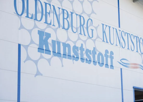 Produktionswerk 1 der Oldenburg Kunststoff-Technik GmbH in Chieming/Bayern Hallenfassade mit blauem Schriftzug „OLDENBURG KUNSTSTOFF“ und grafischen Kreisflächen.