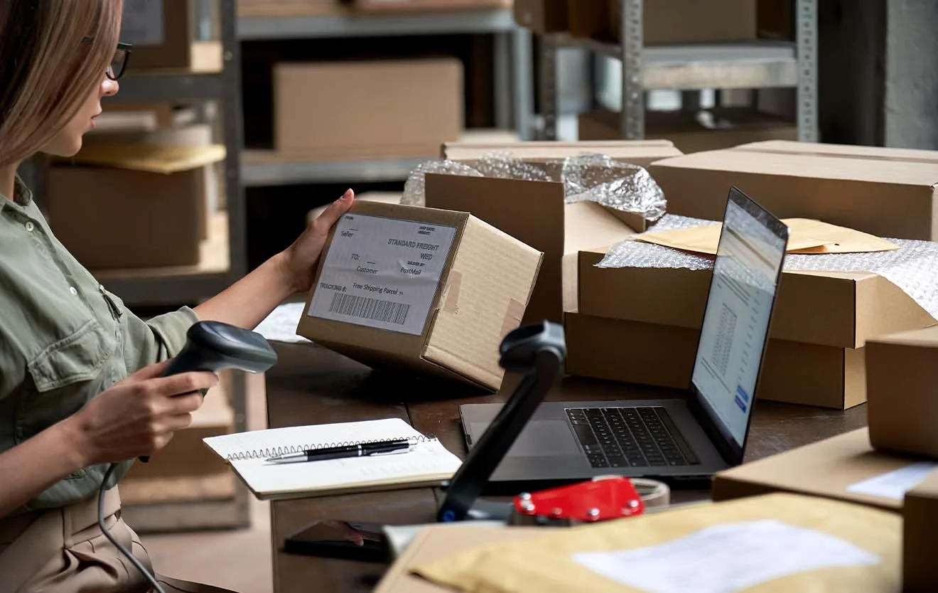 An employee scans a parcel and works on a laptop between boxes.