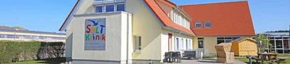 SyltKlinik der Deutschen Kinderkrebsstiftung mit Außenbereich und Strandkorb SyltKlinik der Deutschen Kinderkrebsstiftung mit rotem Dach, Terrasse und Strandkorb im Garten.